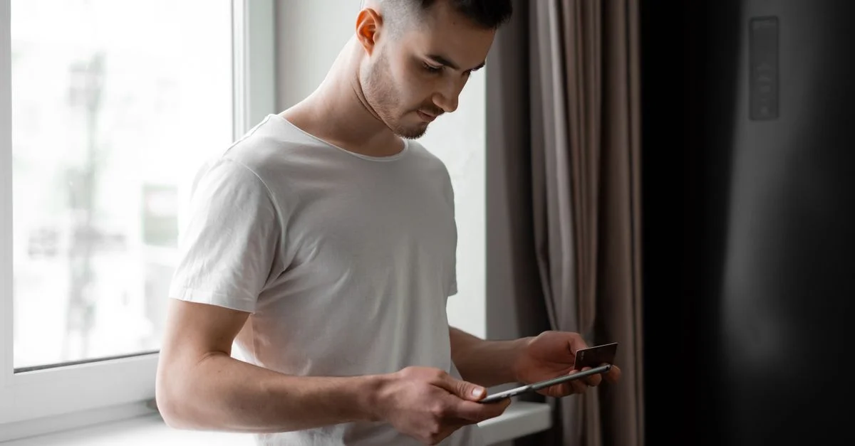 Jeune homme debout près d'une fenêtre, utilisant une tablette et une carte de crédit pour des achats en ligne à l'intérieur.