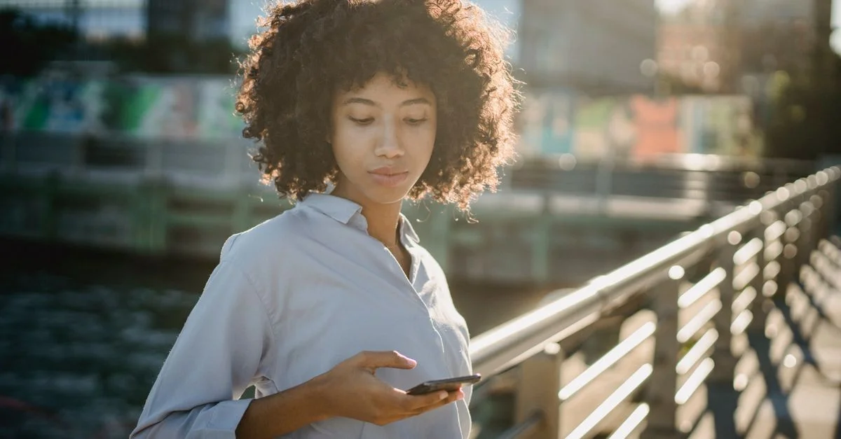 Portrait d'une jeune femme aux cheveux bouclés utilisant son smartphone sur un pont urbain pendant la journée.
