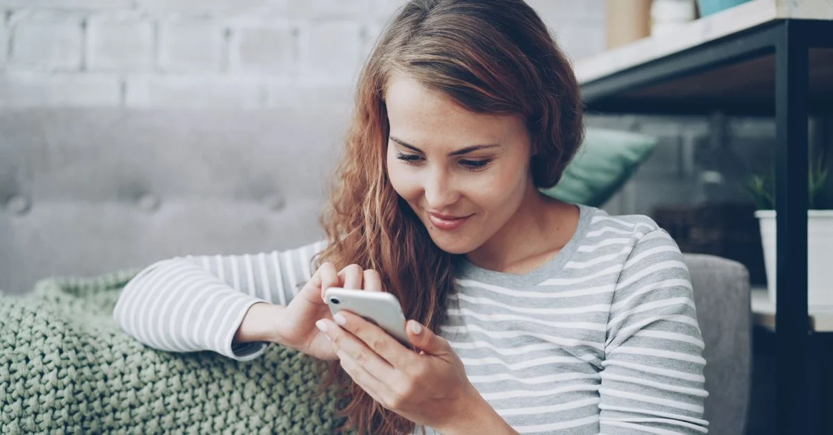 Jeune femme en tenue décontractée utilisant un smartphone, assise confortablement à l'intérieur.